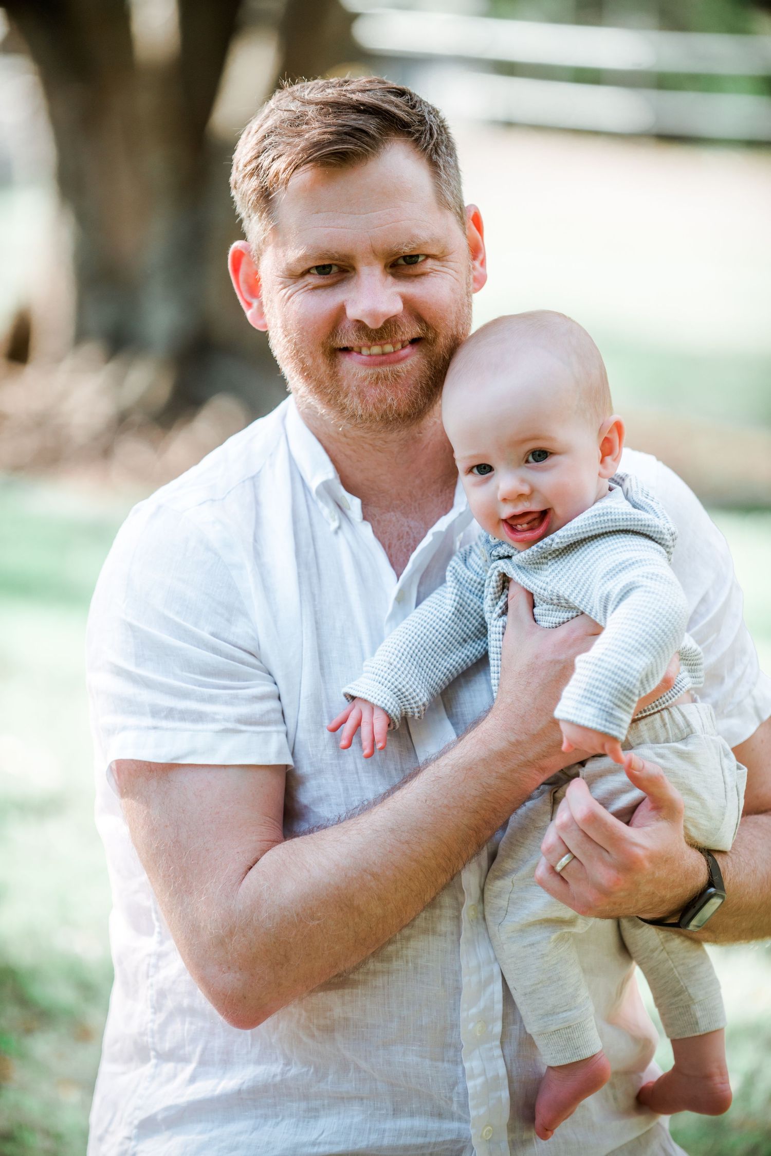 Outdoor portrait of adult holding happy baby in light blue outfit against natural background.