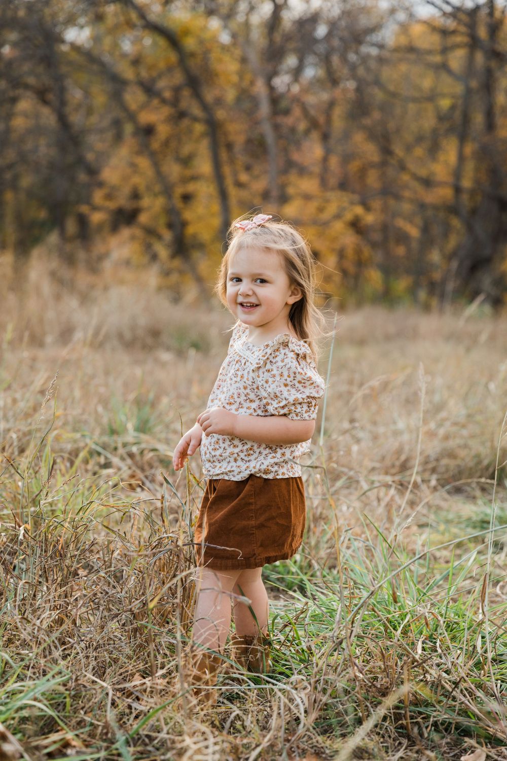 Young child in floral top and brown shorts smiling in autumn field setting.