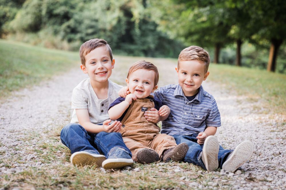 Three siblings in casual attire sitting together on a gravel path surrounded by greenery.