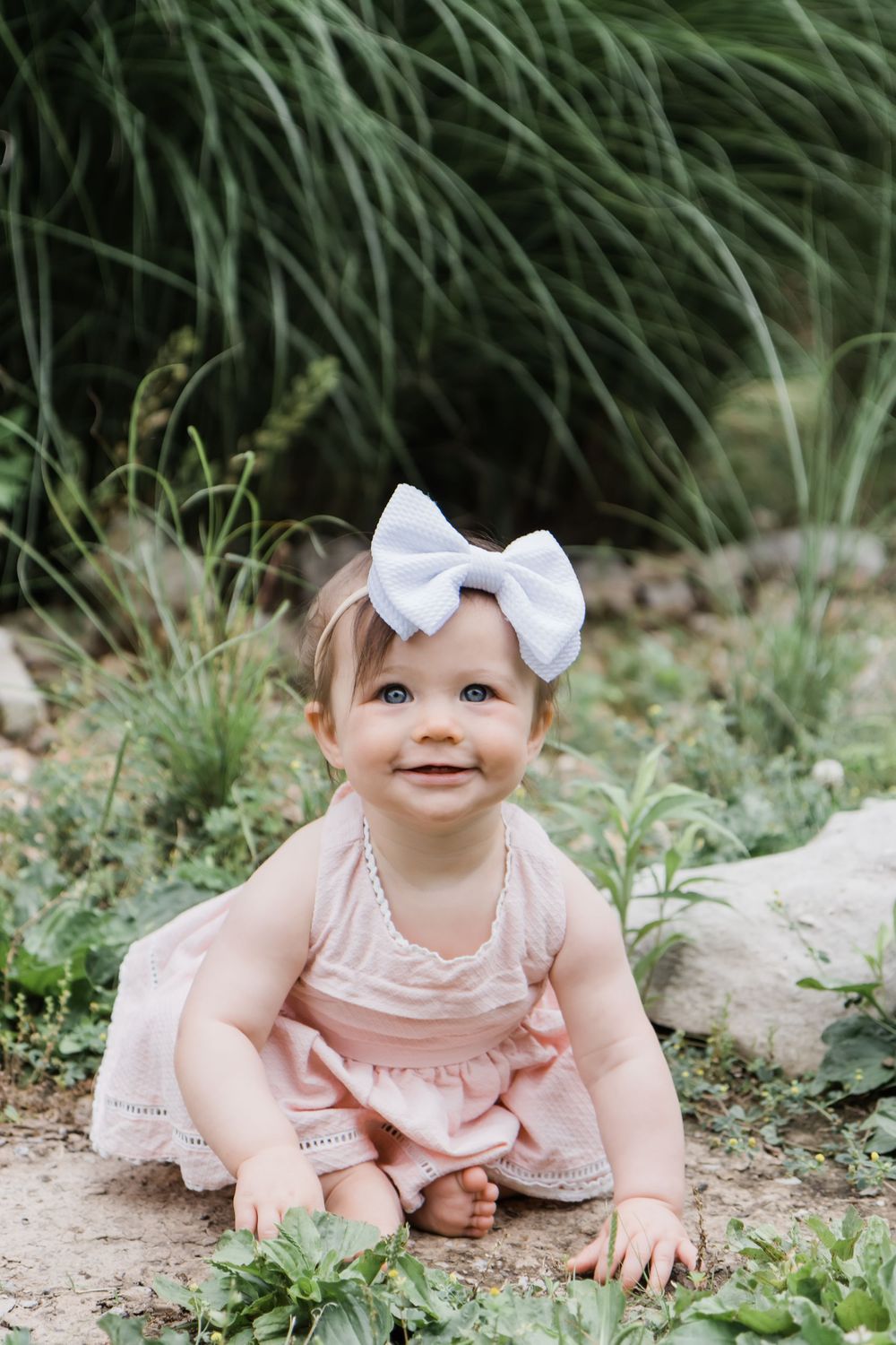 Baby in pink dress and white bow sitting among garden greenery.