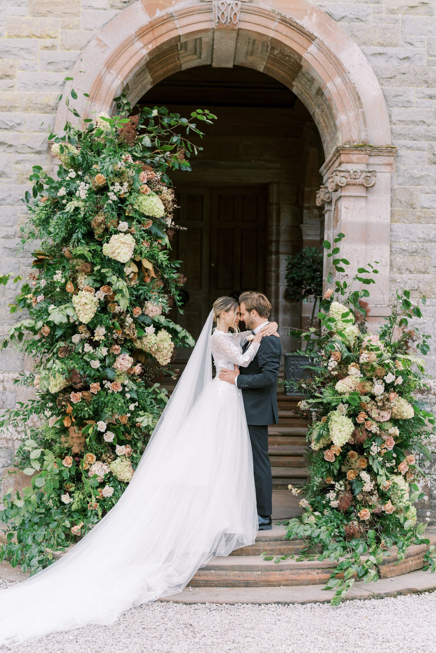 Wedding couple embraces under stone archway adorned with climbing peach and cream roses.