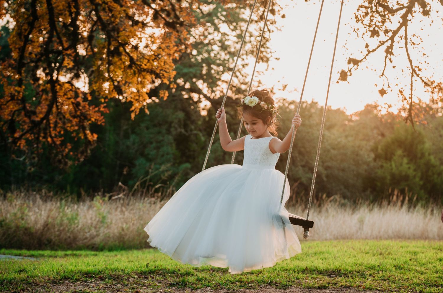 A white tulle dress catches the golden sunset light as someone swings beneath autumn trees in a peaceful garden setting.