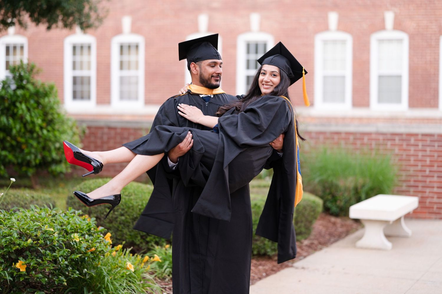 Two graduates in black caps and gowns celebrate joyfully outside a brick building.