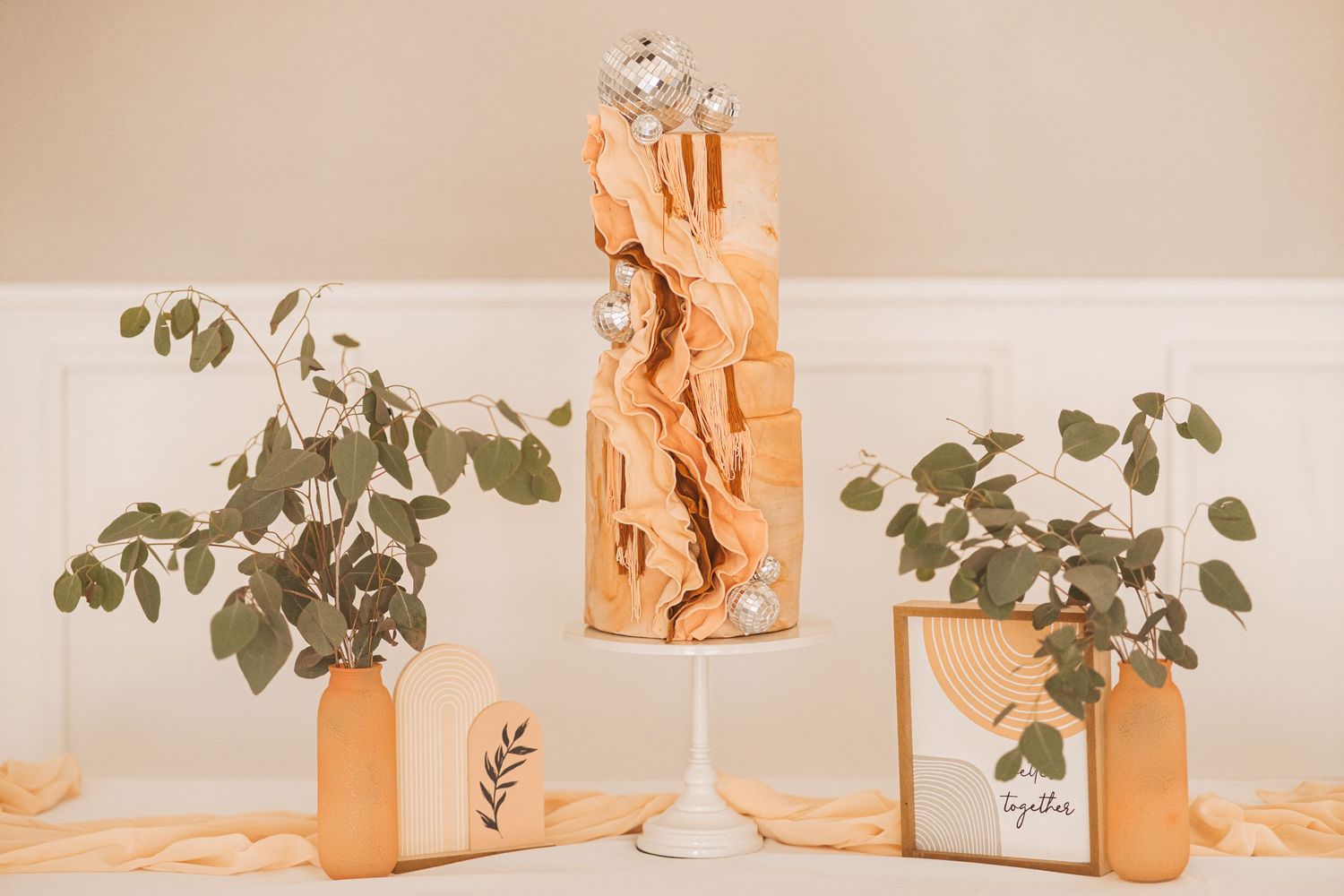 Modern wedding cake display with eucalyptus leaves and warm lighting.