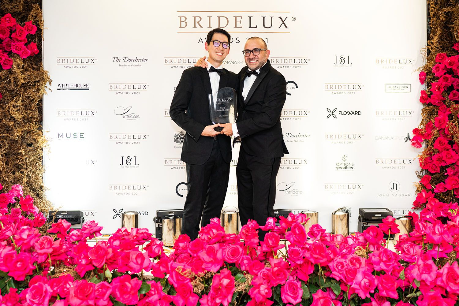 Event attendees pose with award against backdrop of vibrant pink flowers at formal ceremony.
