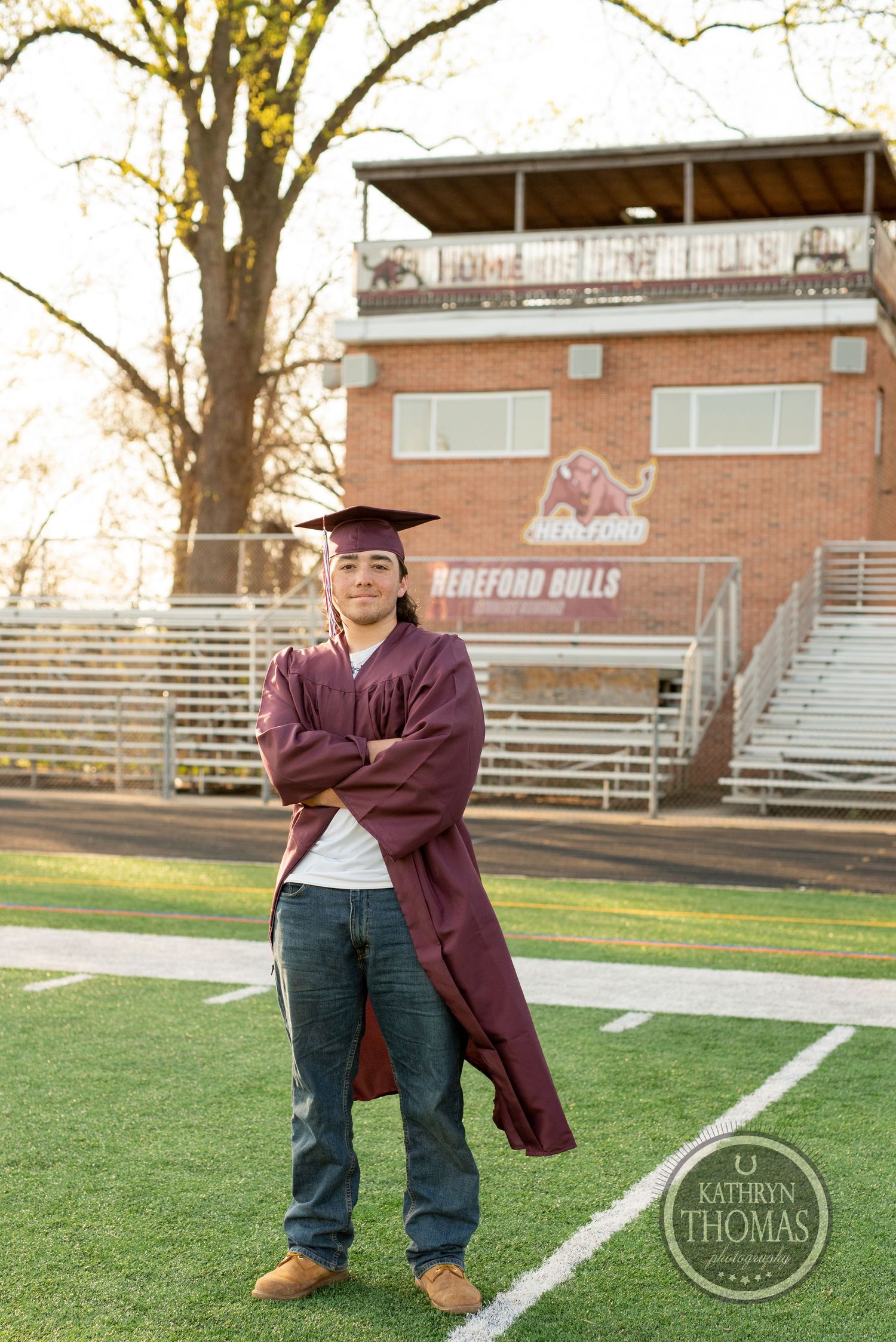 Graduate in maroon graduation gown poses on football field with stadium bleachers in background.
