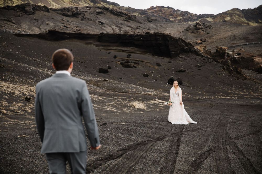 Wedding couple stands apart on volcanic terrain with mountains and cloudy sky.