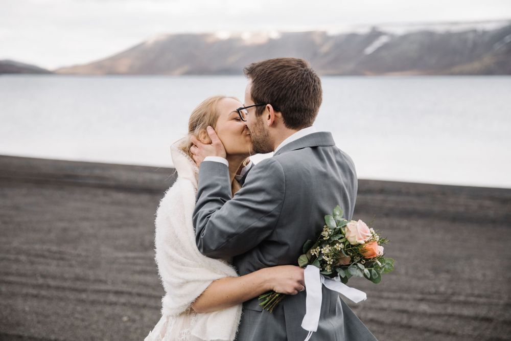 Newlyweds embrace while holding a bouquet against a dramatic mountain and lake landscape.