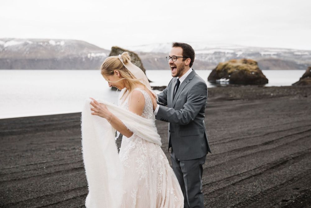 A couple shares a joyful moment on a black sand beach with snow-capped mountains in the background.