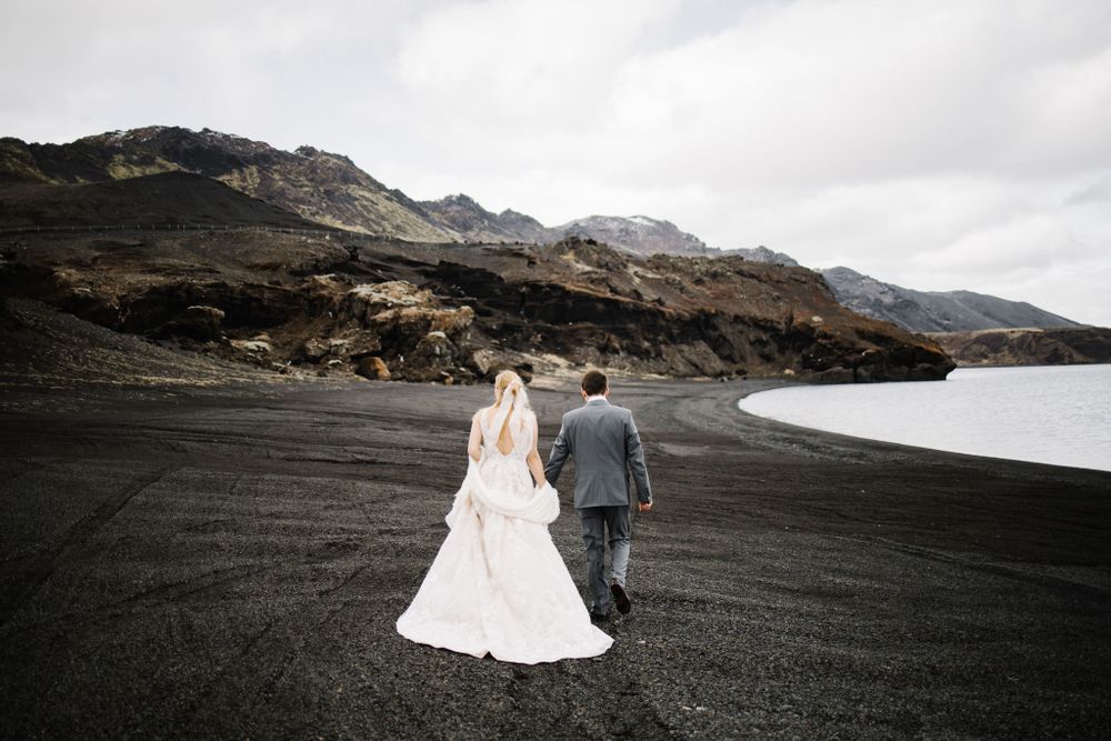 Wedding couple walking together on black volcanic sand with mountains and ocean in view.