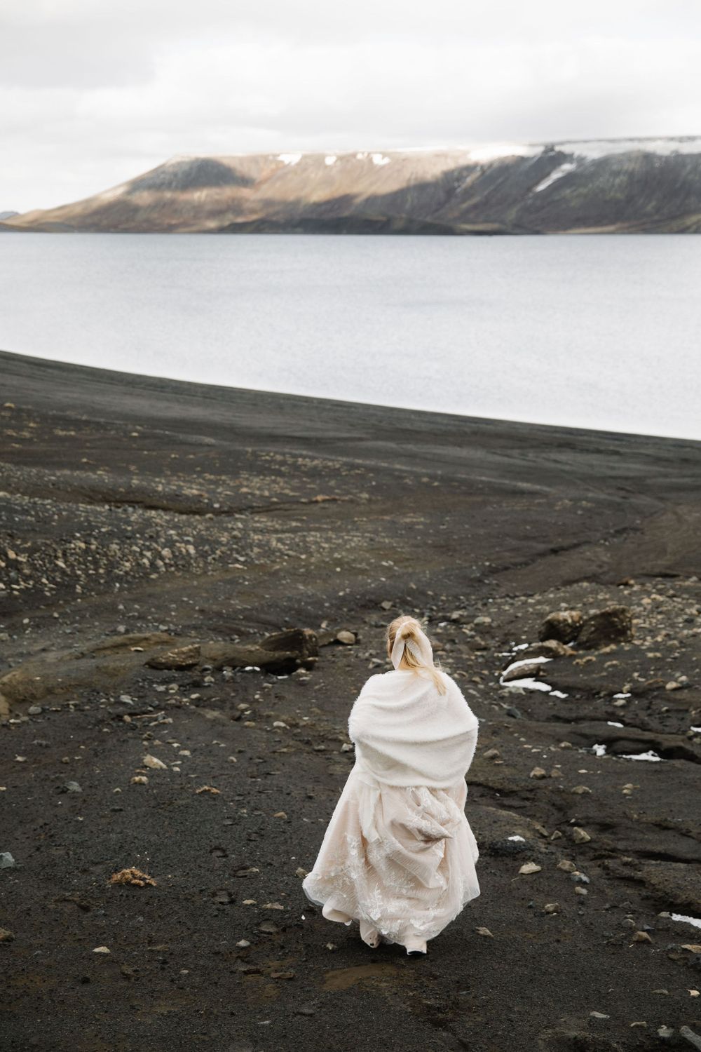 Person in white wedding dress walks on black sand beach with mountain range and lake in background.