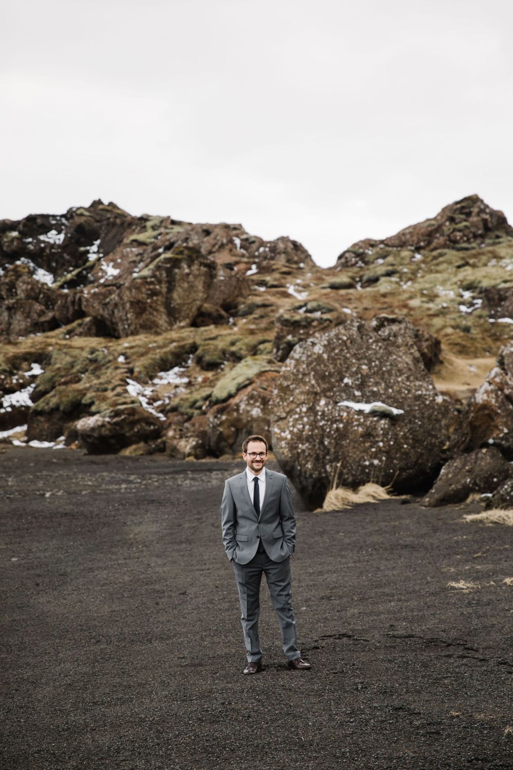 Person in formal suit stands on black volcanic sand with rocky terrain and snow patches in background.