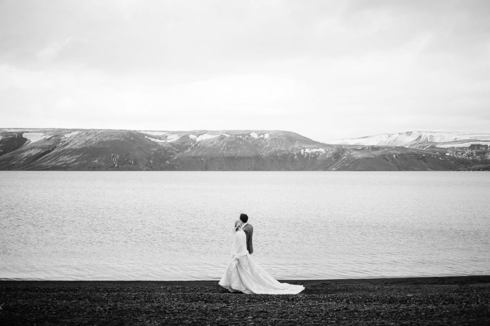 Black and white silhouette of a bride standing by a lake with snow-capped mountains in the distance.