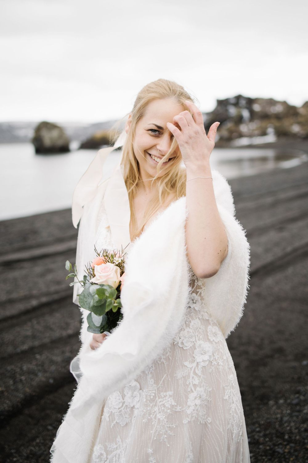 Joyful bridal portrait on a black sand beach with white fur wrap and pink floral bouquet.