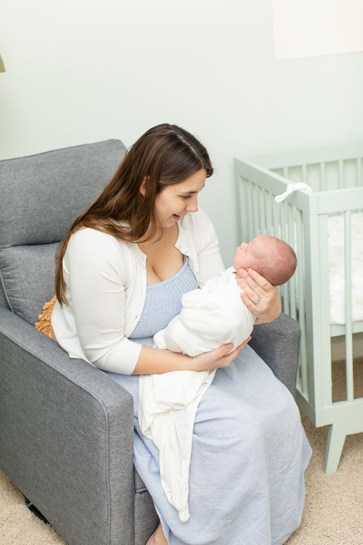 Woman in white cardigan holding newborn baby while sitting in gray nursery chair.