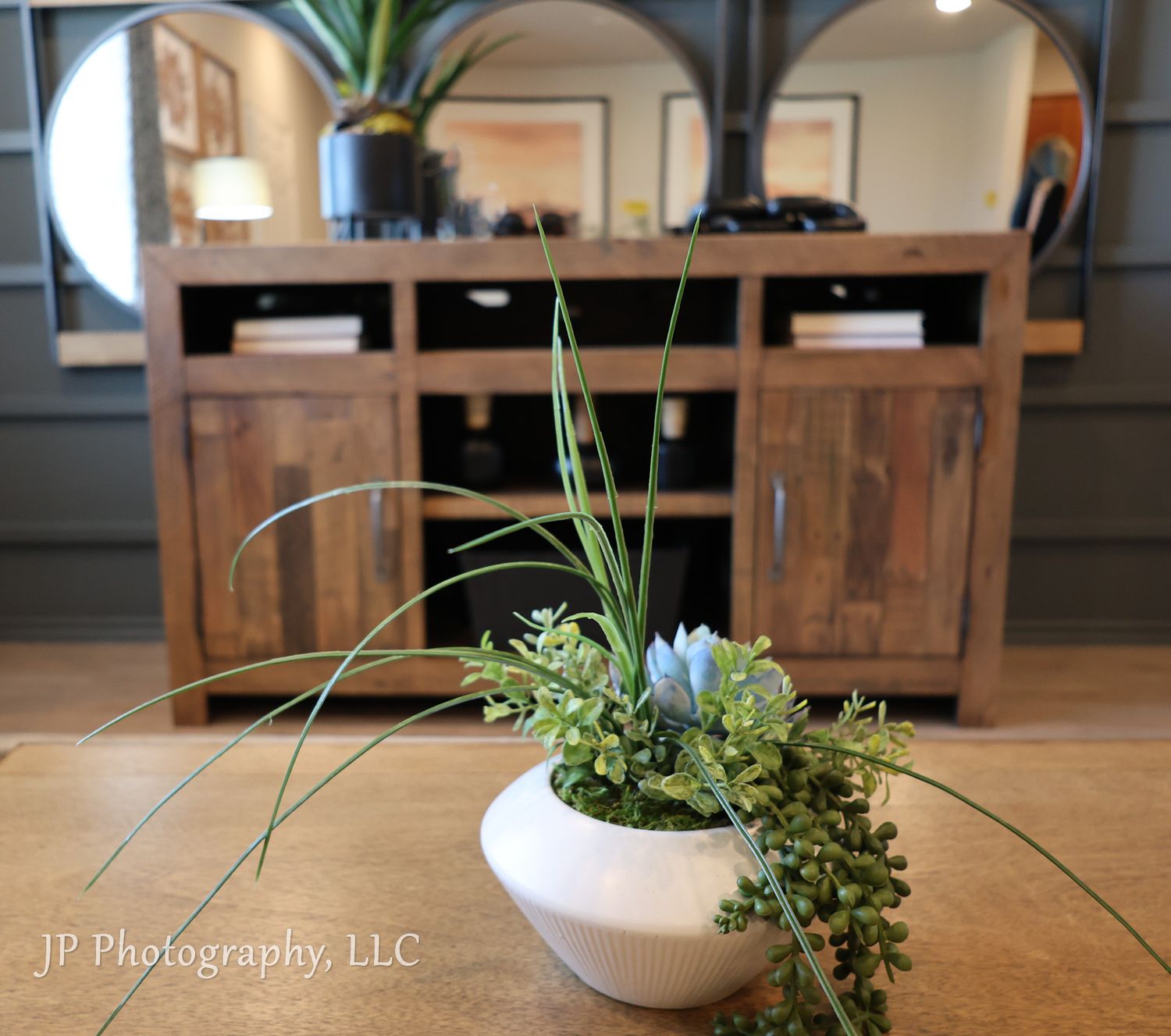 Small white bowl with green plants and grass arranged on wooden table with cabinet in background.