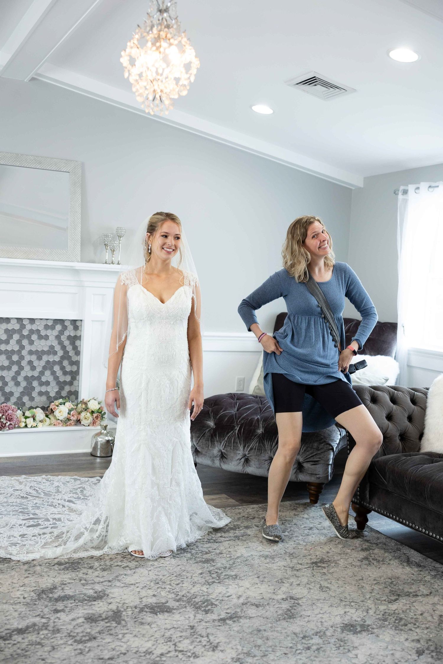 Two figures sharing a joyful moment in an elegant living room with one in a wedding dress beside a grey sofa.
