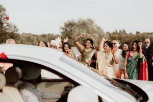 Wedding guests in colorful attire gather excitedly around a car during a celebration.
