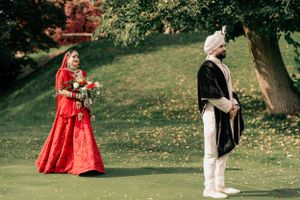 A couple in traditional wedding attire walking in a garden setting with red lehenga and cream sherwani.