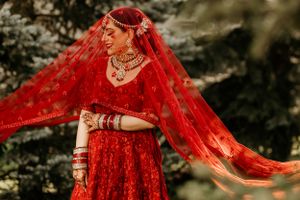 Artistic shot of bride in flowing red lehenga and veil against dark forest backdrop.