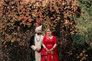 Traditional Indian wedding couple in red and cream attire posing against autumn foliage.
