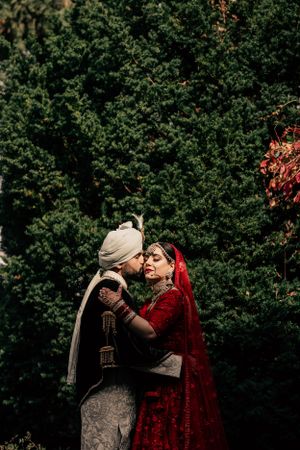 Romantic moment between newlyweds in traditional Indian wedding attire against green foliage.