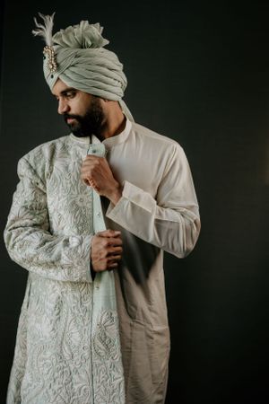 Traditional embroidered sherwani being adjusted against dark background in elegant pose.