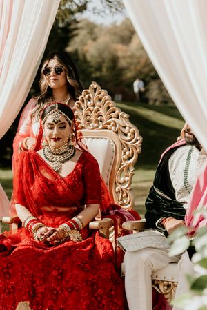 Traditional South Asian bride in red lehenga seated on ornate throne during outdoor ceremony.