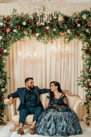 Couple sharing intimate moment on vintage loveseat under floral wedding arch.