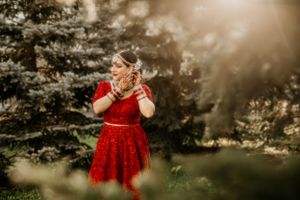 Ethereal portrait of bride in red dress among sunlit evergreen trees.