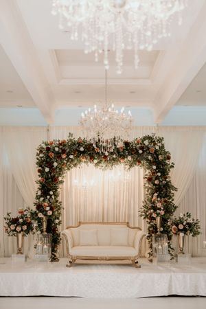 Elegant wedding stage decorated with floral arch, crystal chandeliers and cream-colored loveseat.