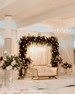 Luxurious wedding reception setup with floral arch and crystal chandeliers over white loveseat.