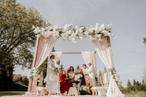 Wedding ceremony under a pink and white floral arch with gathered guests on a sunny day.