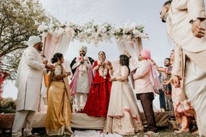 Wedding party celebrating during traditional Indian ceremony under pink and white floral arch.