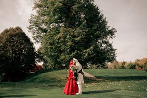 A couple in wedding attire stands on a lush green lawn beneath tall trees during a romantic outdoor photo session.