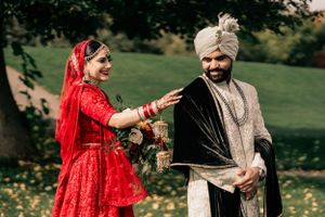 Traditional Indian wedding couple in red and cream attire sharing a playful moment outdoors.