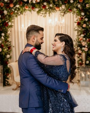 Couple in navy and sparkly formal wear embrace under a floral arch at their reception.
