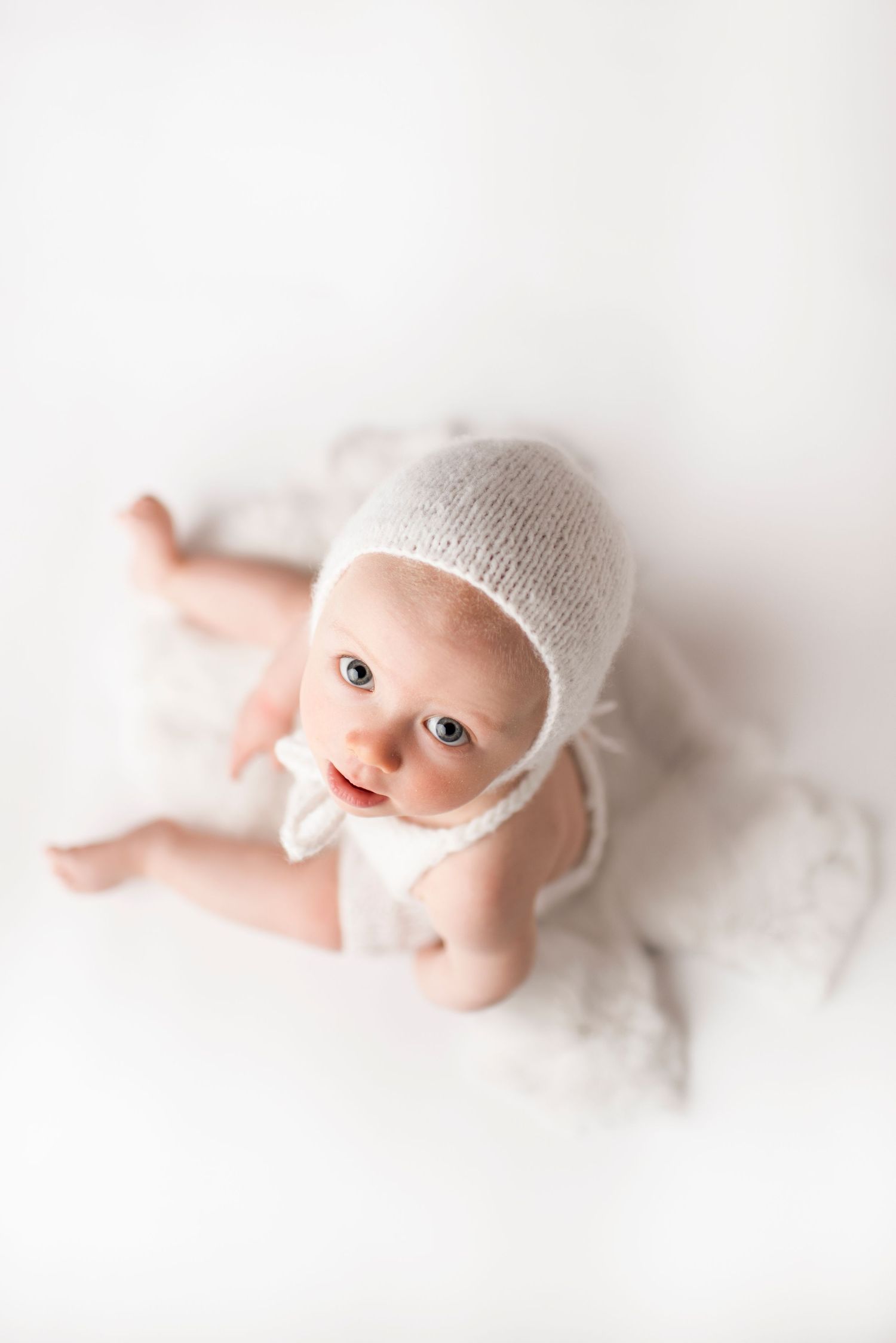 A baby in white bonnet and outfit poses on white backdrop during studio photography session.