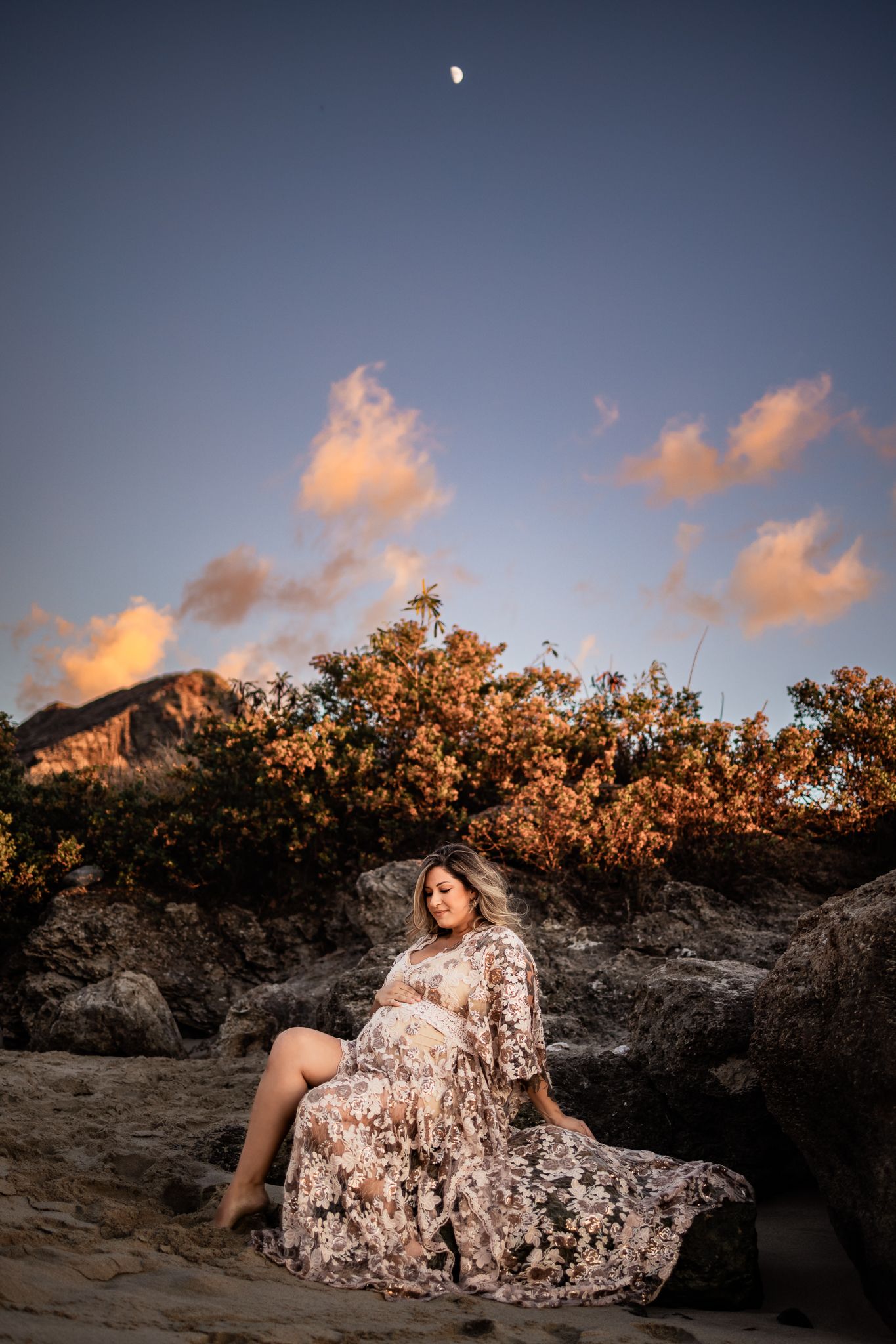 A woman in a floral dress poses on rocks at sunset with a mountain and moon in the background.