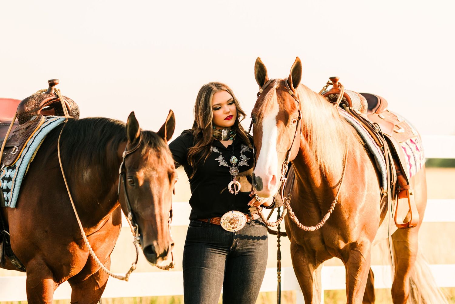 Woman stands between two saddled horses at sunset.
