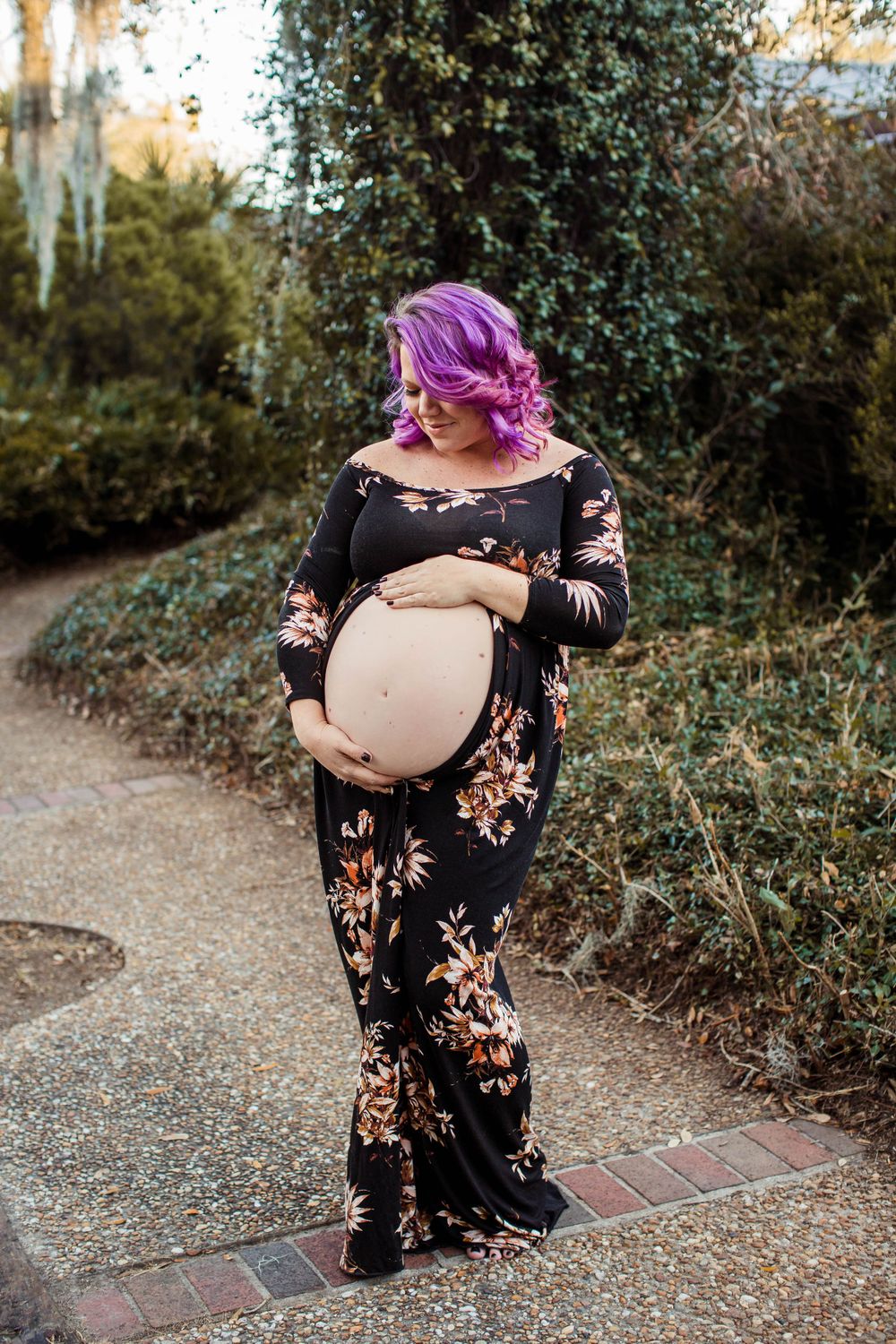 A maternity portrait of someone in a floral dress with vibrant purple hair on a garden path.