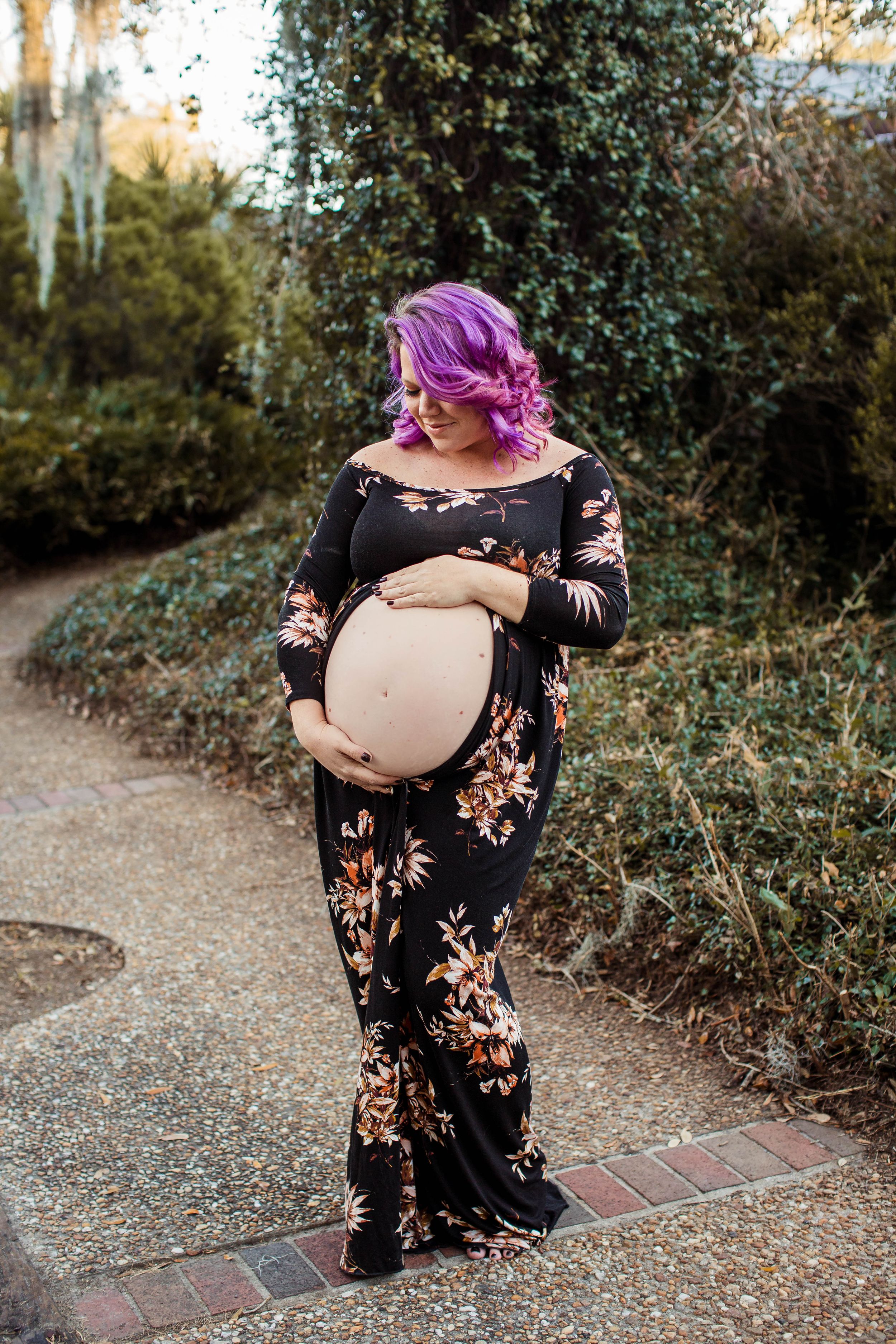 A maternity portrait of someone in a floral dress with vibrant purple hair on a garden path.