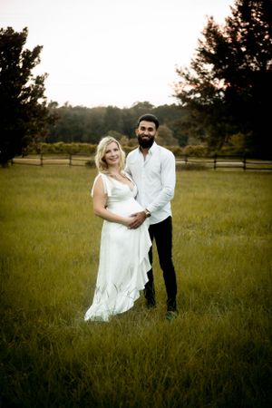 Expecting couple stands together in rustic field at sunset wearing white dress and casual attire.