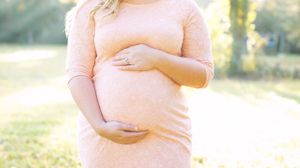 Gentle hands cradling a pregnant belly wearing a soft pink sweater in golden sunlight.
