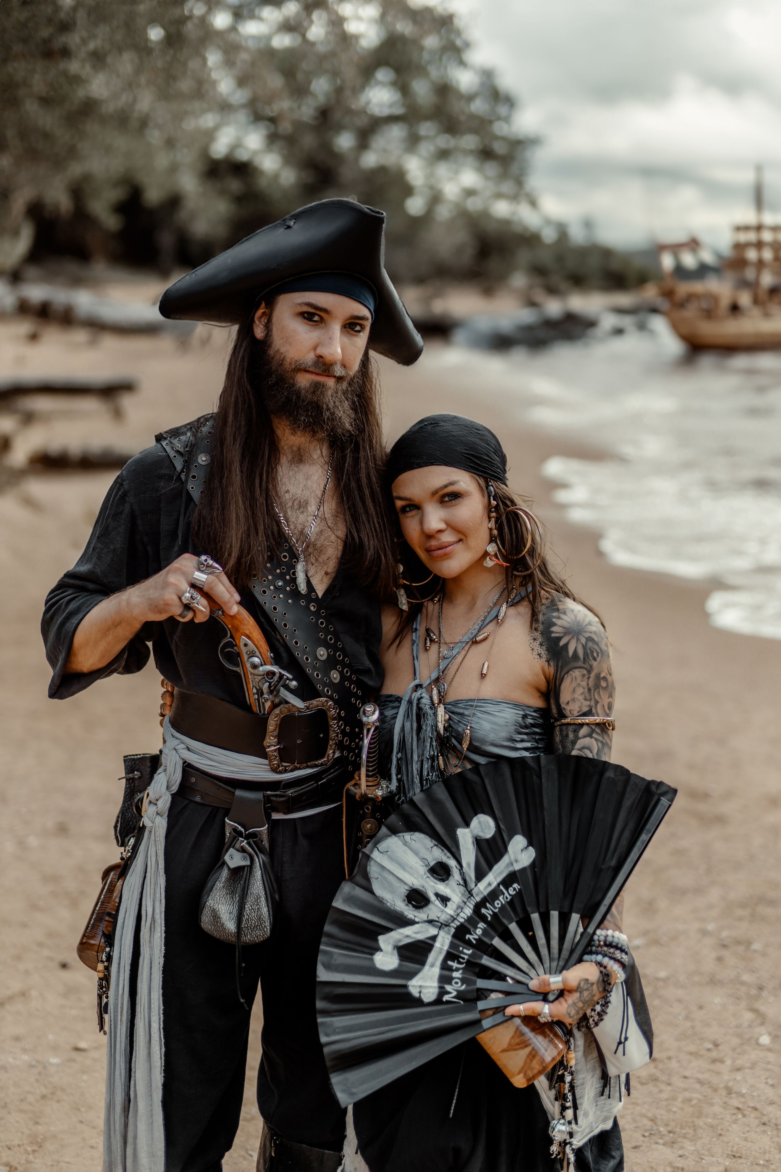 A couple in pirate costumes pose on a beach with a skull fan and vintage ship in the background.