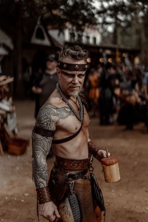 A shirtless warrior with tribal tattoos and leather armor stands in a medieval festival setting.