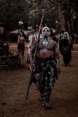 A tribal warrior with face paint and body art carries a staff at a renaissance festival.