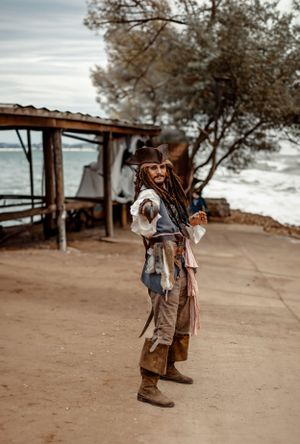 A pirate cosplayer poses on a beach path with boats visible in the background.
