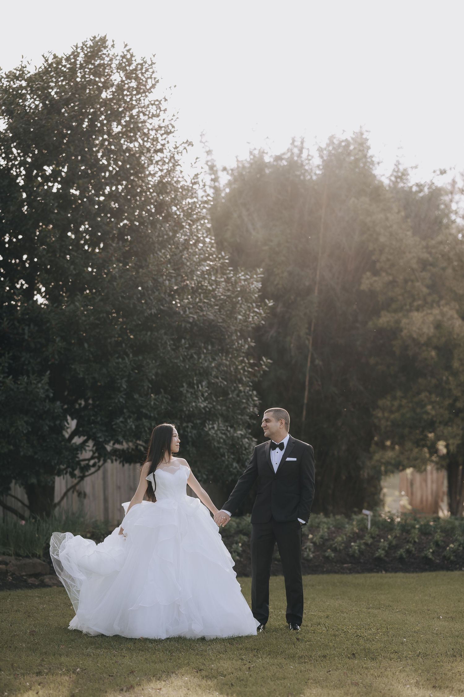 Elegant couple in wedding attire standing together on a sunlit lawn with trees in background.