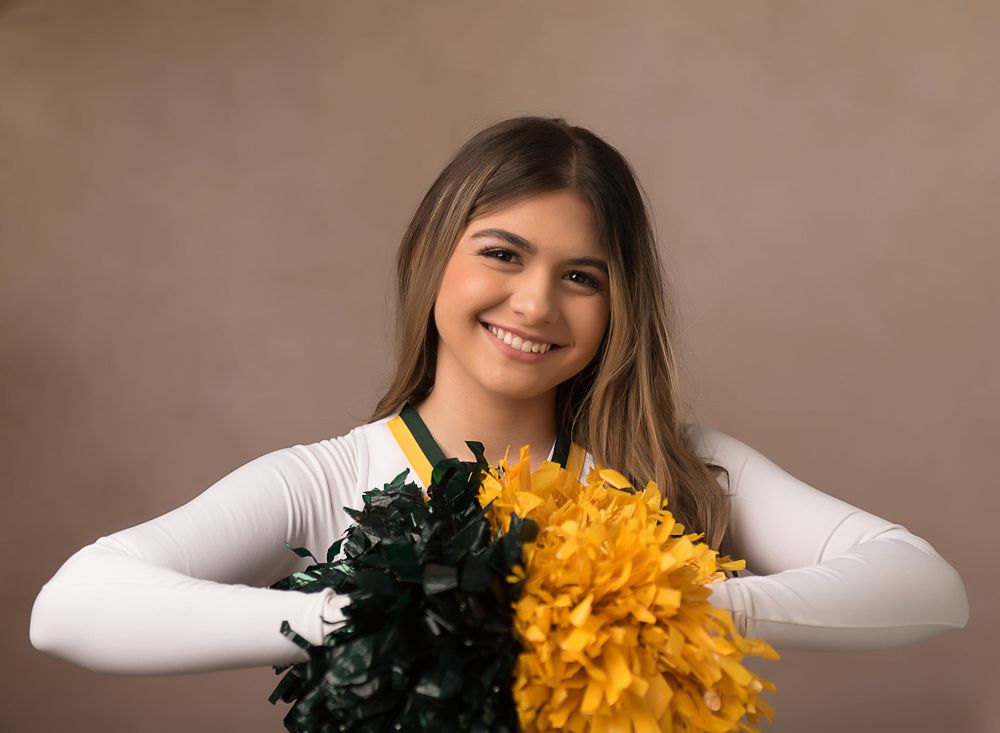 Cheerleader holds green and gold pom poms while posing against neutral background.